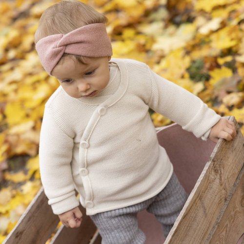 Young girl in wooden cart looking down. The girl is dressed with the disana leggings in the colour grey, the disana melange jacket in the colour nature and the disana headband in the colour rosé