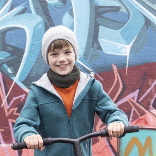 Teenager with the disana tube scarf in the two-coloured melange olive anthracite rides a scooter in front of a graffiti wall and smiles