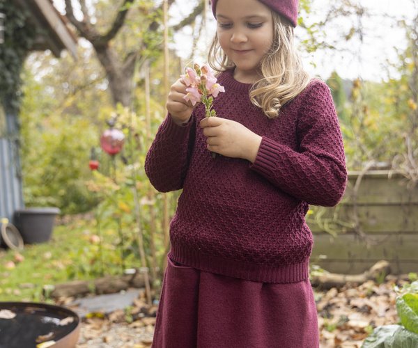 Blonde girl with disana boiled wool skirt in cassis, aran sweater in cassis and headband in cassis holds pink flowers in her hands